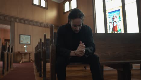 young, anxious man in black suit sitting in pew in church praying in worship in cinematic slow motion