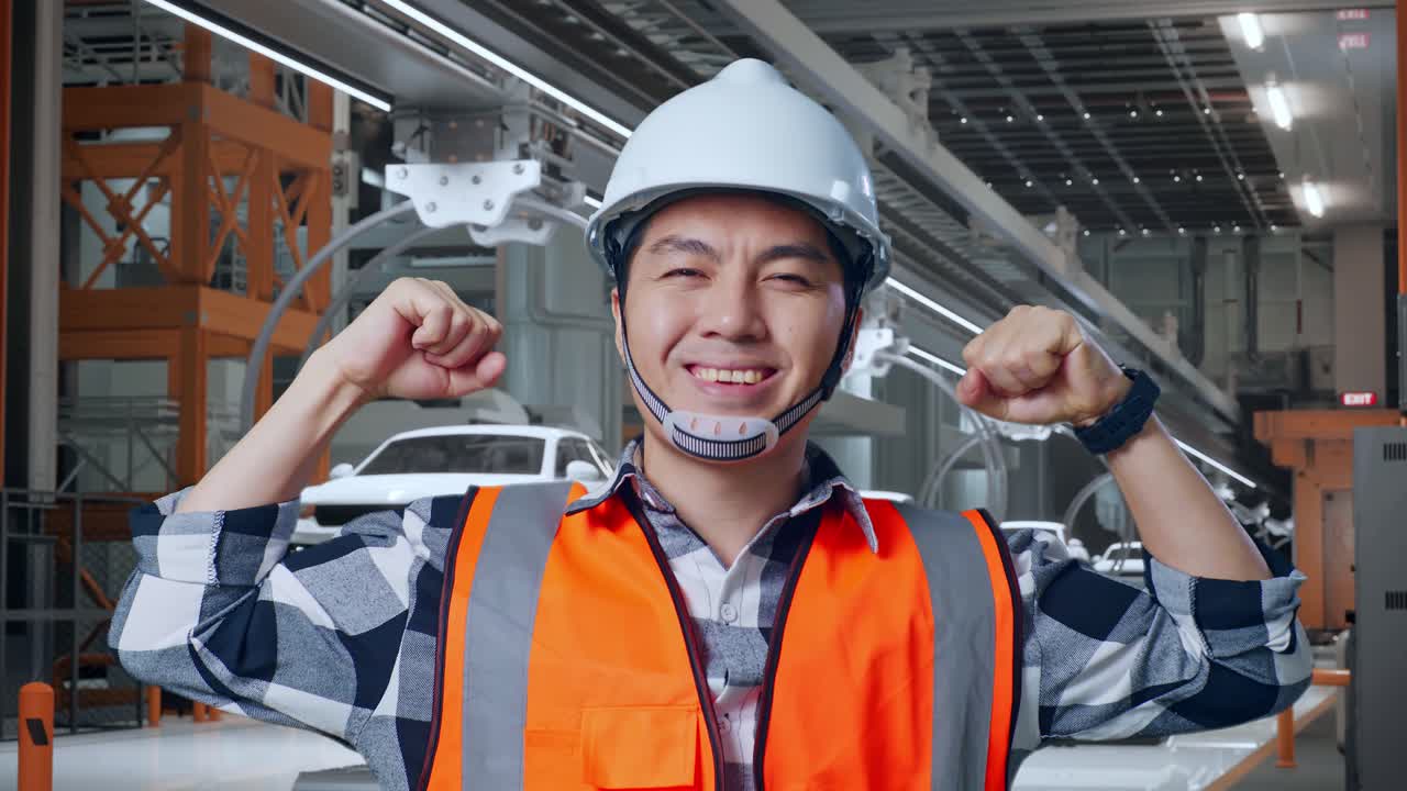 안전 헬을 착용한 아시아 남성 엔지니어가 ev 자동차 공장에서 서서 카메라에 미소 짓고 이를 구부리고 있습니다.