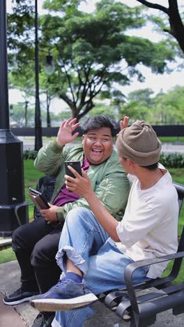 two college students using smartphone and giving high five to each other while sitting on bench at campus park