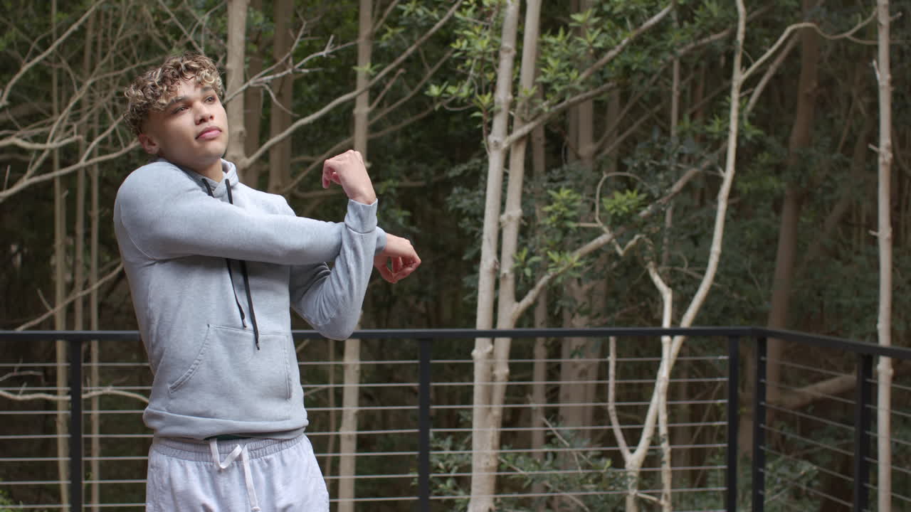 Young man stretching arms outdoors, wearing gray hoodie, surrounded by trees, copy space