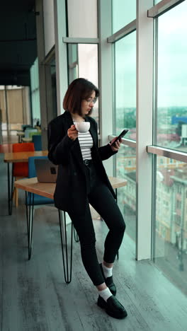 caucasian girl leaning on the desk holding a cup. full-length portrait of a relaxed lady on a break from work using her phone. vertical video.