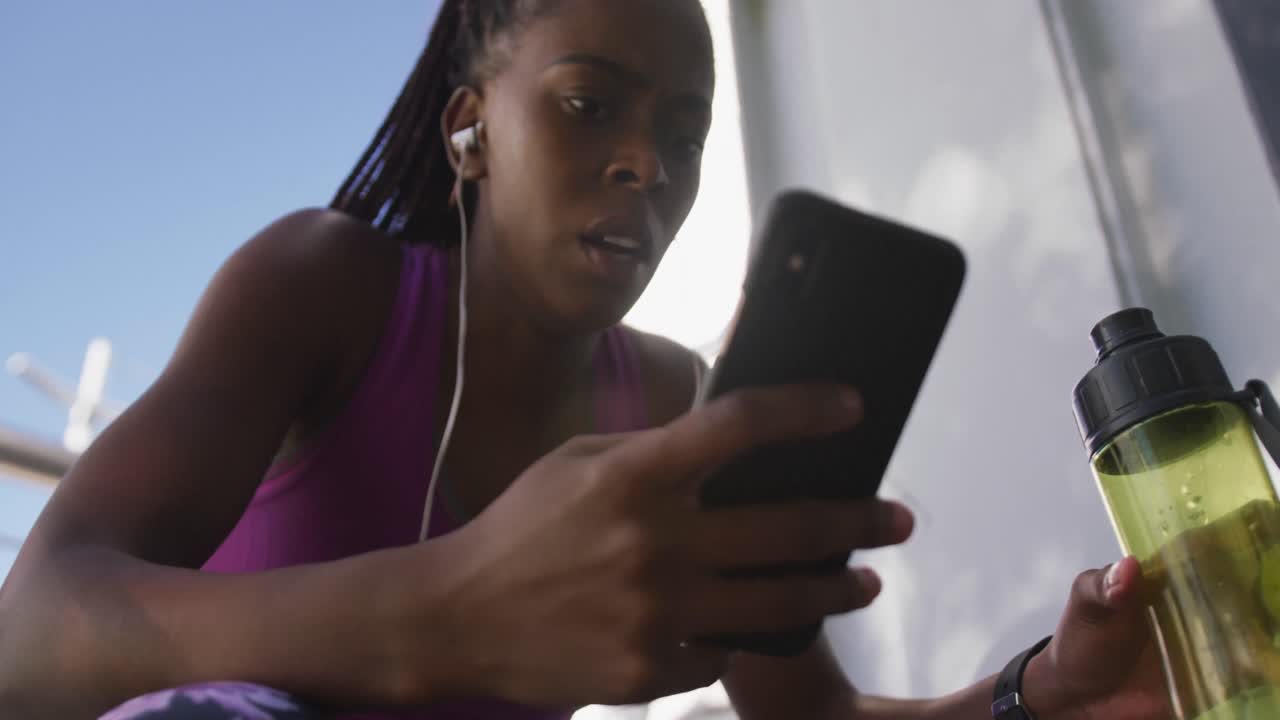African american woman smiling while using smartphone sitting on the stairs of the city bridge