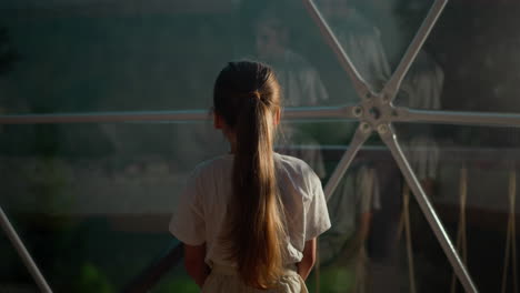 child touches metal carcass of dome at glamping. little girl studies spheric panoramic window with frame in hotel. kid rests in luxury camp