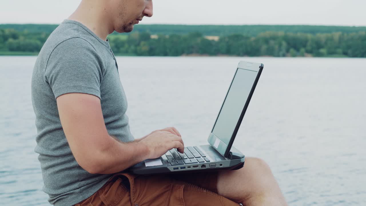 Handsome man typing on a laptop sitting over the flowing river and enjoys the nature in summer. Young male combines his work with a rest outdoors. Close-up