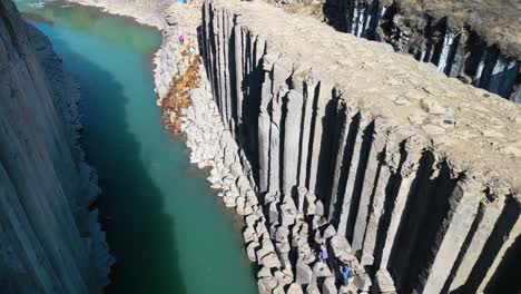 Aerial-drone-shot-of-tourist-walking-around-scenic-icelandic-studlagil-canyon-with-river-flowing-into-basaltic-rock-formation-in-Iceland-on-a-sunny-day