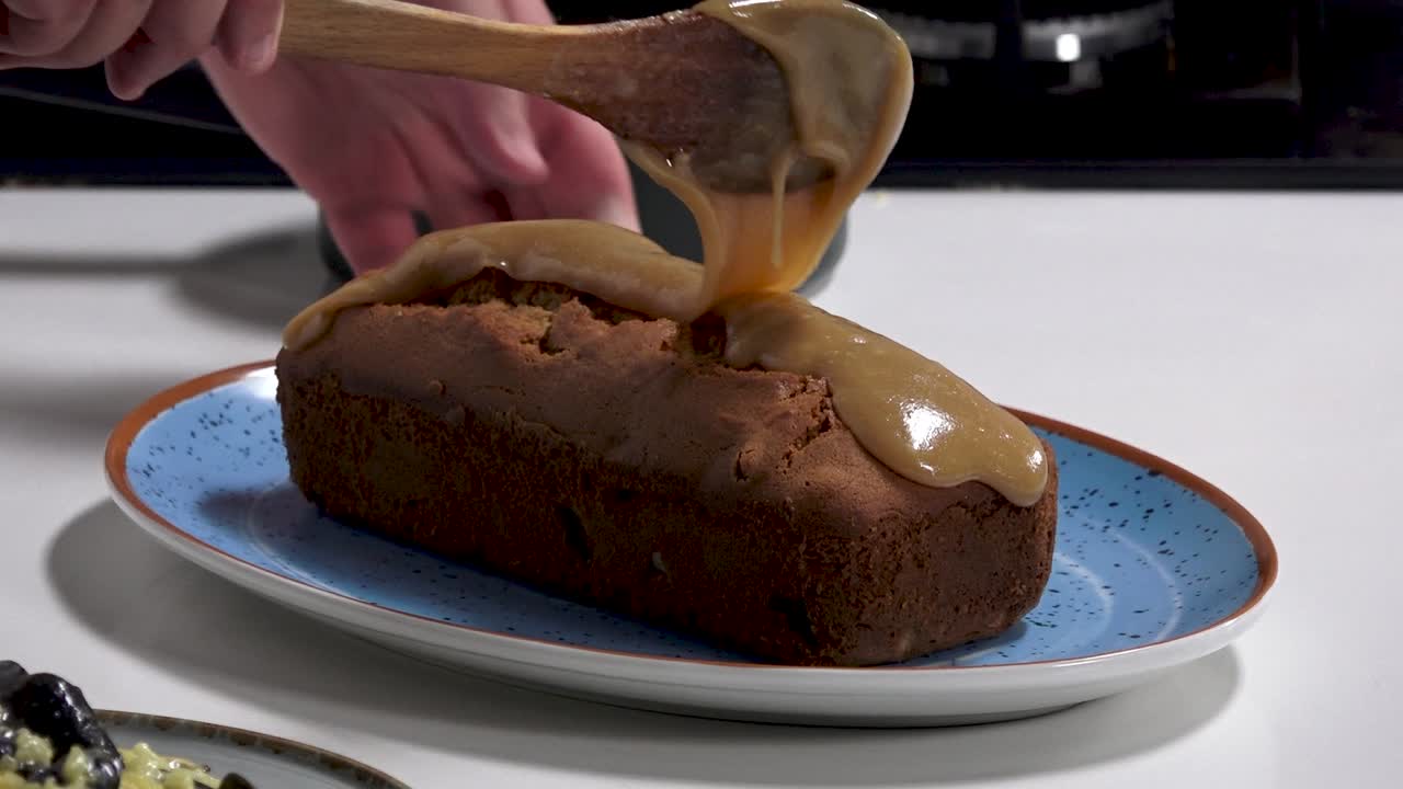 A freshly baked loaf cake is elegantly topped with warm caramel sauce. The process takes place in a welcoming kitchen, enhancing the delightful aroma of baking
