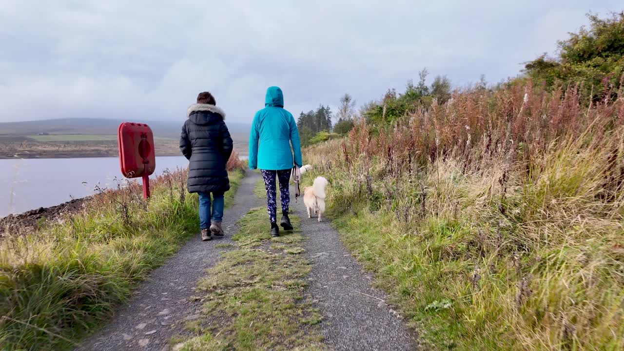 People and dog walking along a rural path beside Burnhope Reservoir on a cloudy day