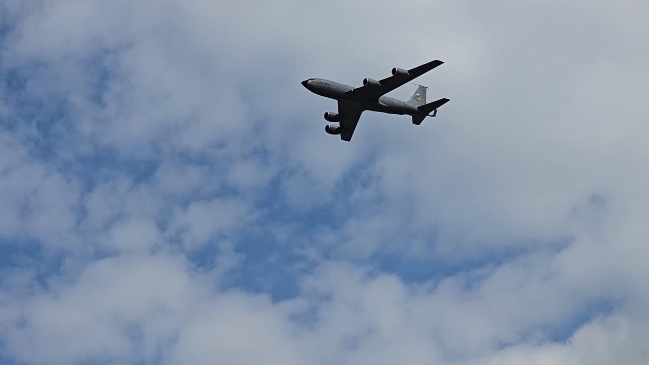 A large military aircraft soars through a bright blue sky with scattered clouds, showcasing its impressive design and power.