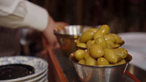 Close-up-on-bronze-metal-cup-filled-with-green-olives,-chef-fills-another-cup-in-background