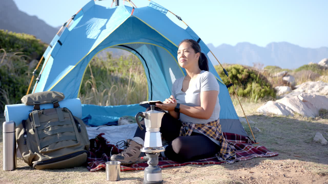 Camping in mountains, woman reading book near tent with camping gear