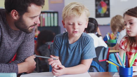 Elementary-School-Teacher-Giving-Male-Pupil-One-To-One-Support-In-Classroom