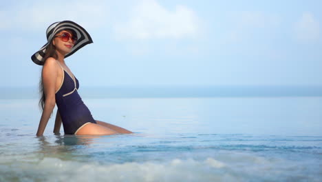 Eine-Hübsche-Junge-Frau-In-Einem-Einteiligen-Badeanzug-Und-Einem-Schlaffen-Sonnenhut-Sitzt-Am-Rand-Eines-überlaufpools-Und-Blickt-Auf-Einen-Ozeanhintergrund