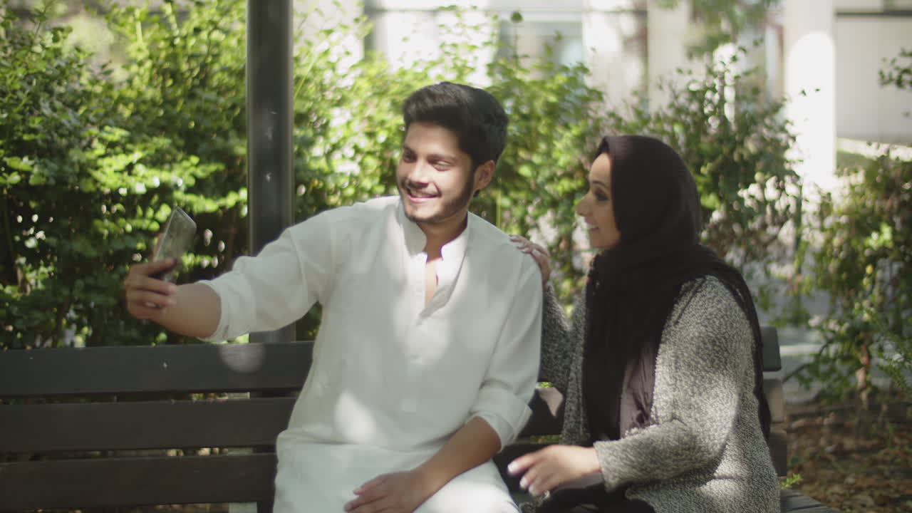 una joven pareja musulmana sentada en un banco, haciendo una llamada telefónica por video.