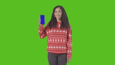 studio portrait of woman wearing christmas jumper holding up mobile phone against green screen 1