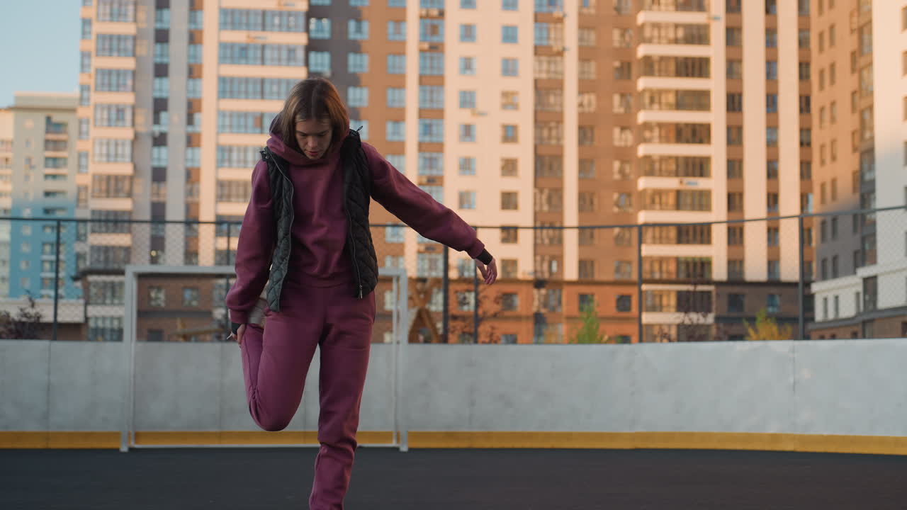 Sportswoman standing on round court outdoors surrounded by modern skyscraper buildings, balancing on one leg while pulling foot to stretch knee with purple tracksuit under evening sun