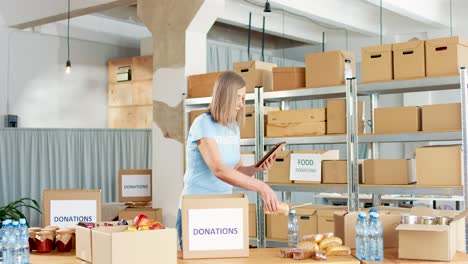 caucasian woman volunteer packing donation boxes with food and water while typing on the tablet in charity warehouse