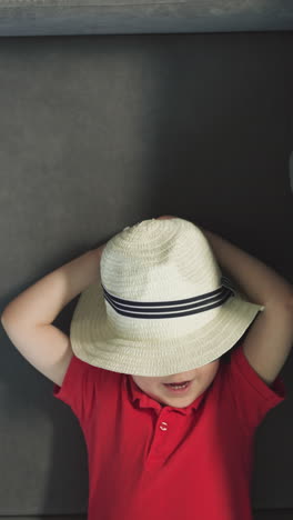 hands of mother pull smiling relaxed toddler in hat on sofa near suitcase. stylish little boy in red t-shirt enjoys resting in room upper close view