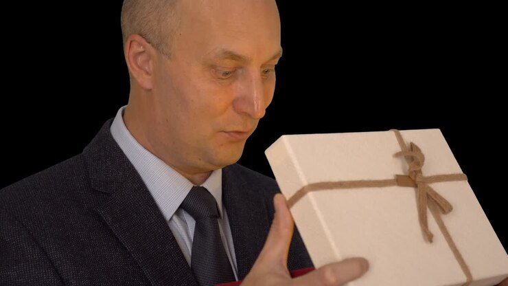 Adult caucasian man in classical formal suit open gift box. Man looking into gift box and spot light at his face. Close up head shot. Alpha channel chroma key transparent background.