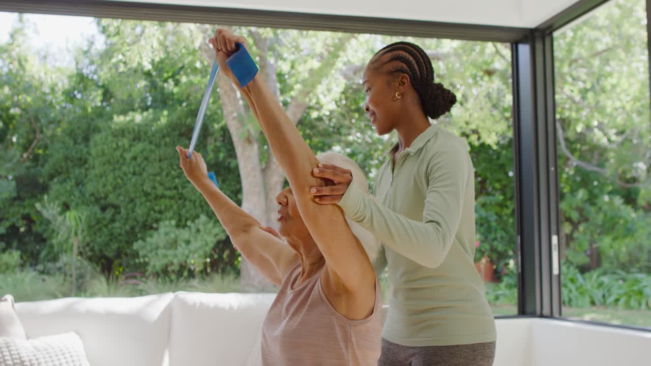 Female carer guiding female senior lifting blue band overhead supporting arm by window for rehab