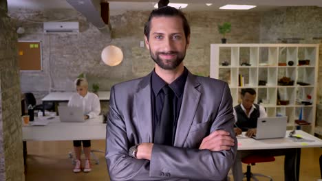 handsome businessman with beard is standing with arms crossed and watching at camera in office, smiling, colleagues are working with laptops, work concept