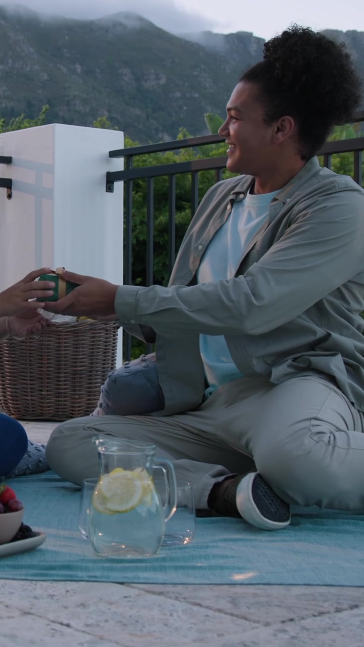 Video verticale: Coppia che si abbraccia su una terrazza, con l'uomo in ginocchio su una coperta che apre una scatola per dare un regalo.