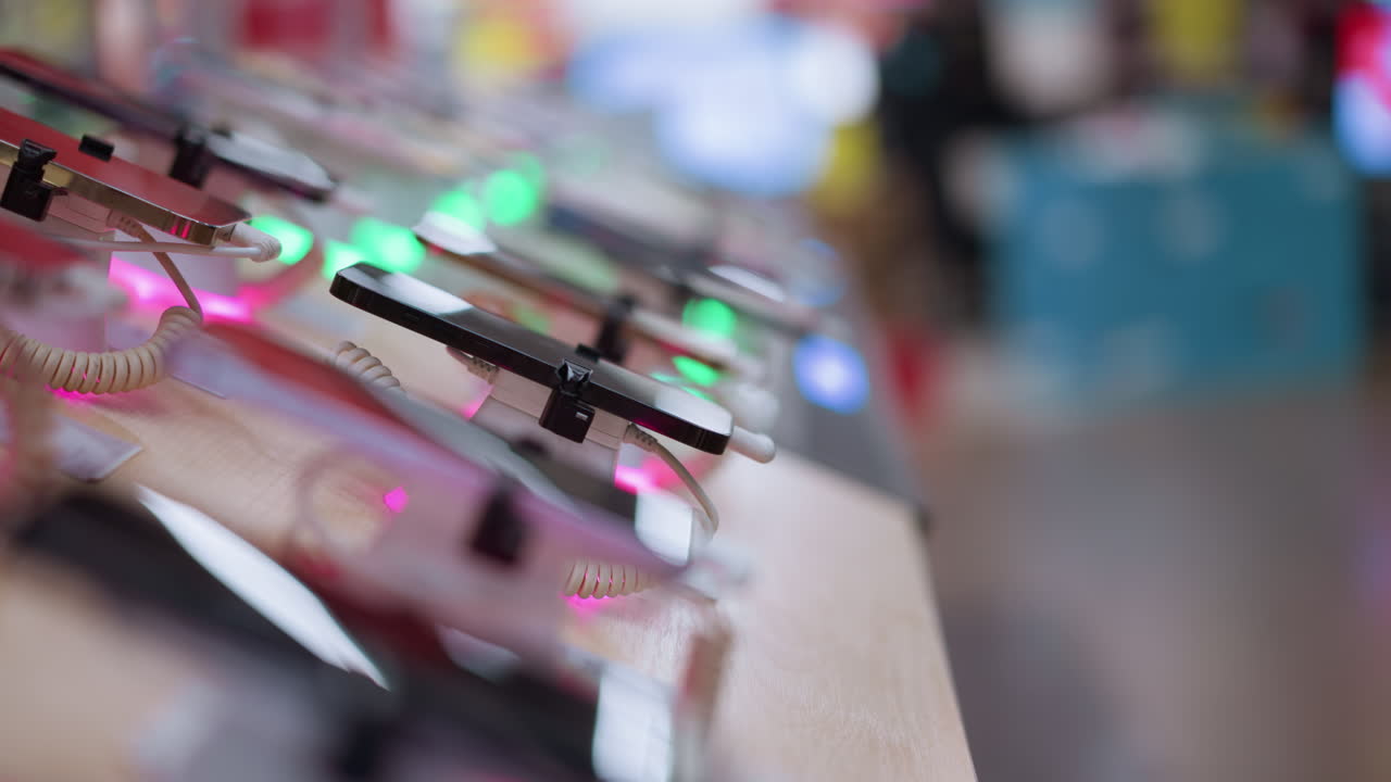 상점에 전시된 스마트폰의 한 줄의 클로즈업 뷰, 각각은 보안 케이블로 연결되어 있으며 다채로운 불빛으로 조명되어 있으며, 배경에는 사람들의 흐릿한 뷰가 있습니다.