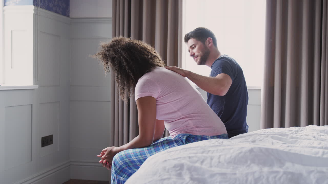 Uomo che conforta una donna vestita che soffre di depressione, seduta sul letto di casa
