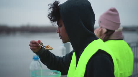 Man in hoodie and neon safety vest eating food from spoon near river while woman in pink jacket and matching vest stands blurred in background, capturing outdoor work lunch break during cold
