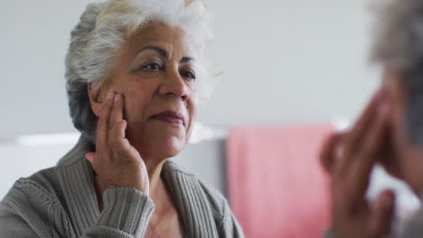 Mujer-Mayor-Afroamericana-Aplicando-Crema-Facial-Mientras-Se-Mira-En-El-Espejo-En-Casa