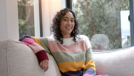 portrait of happy biracial woman sitting on sofa smiling at home, slow motion