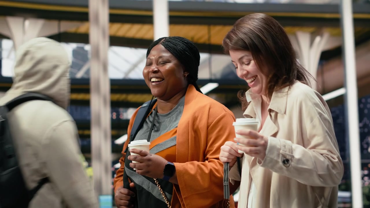 Friends carry drink cups through coach station and discuss travel plans