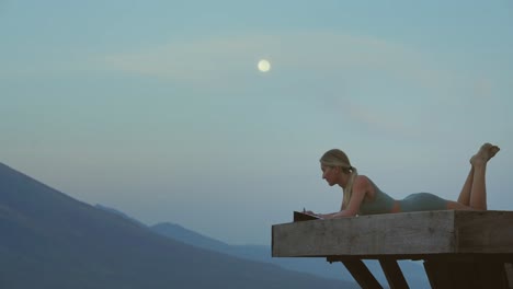 Mujer-Viajera-Escribiendo-En-Diario-Mientras-Se-Tumba-En-Una-Plataforma-De-Madera-Con-Luna-Llena,-Espiritual