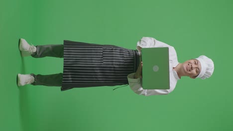 full body of asian man chef using a laptop and smiling to camera while standing in the green screen background studio