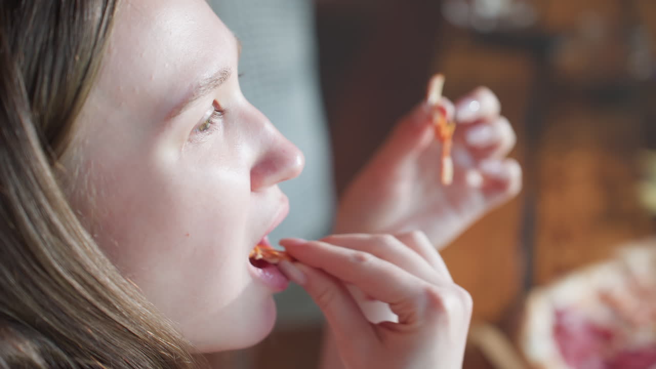 Close up of woman enjoying slice of pizza, holding piece with hands, slight profile view. Another person is visible in the background, adding to casual dining atmosphere