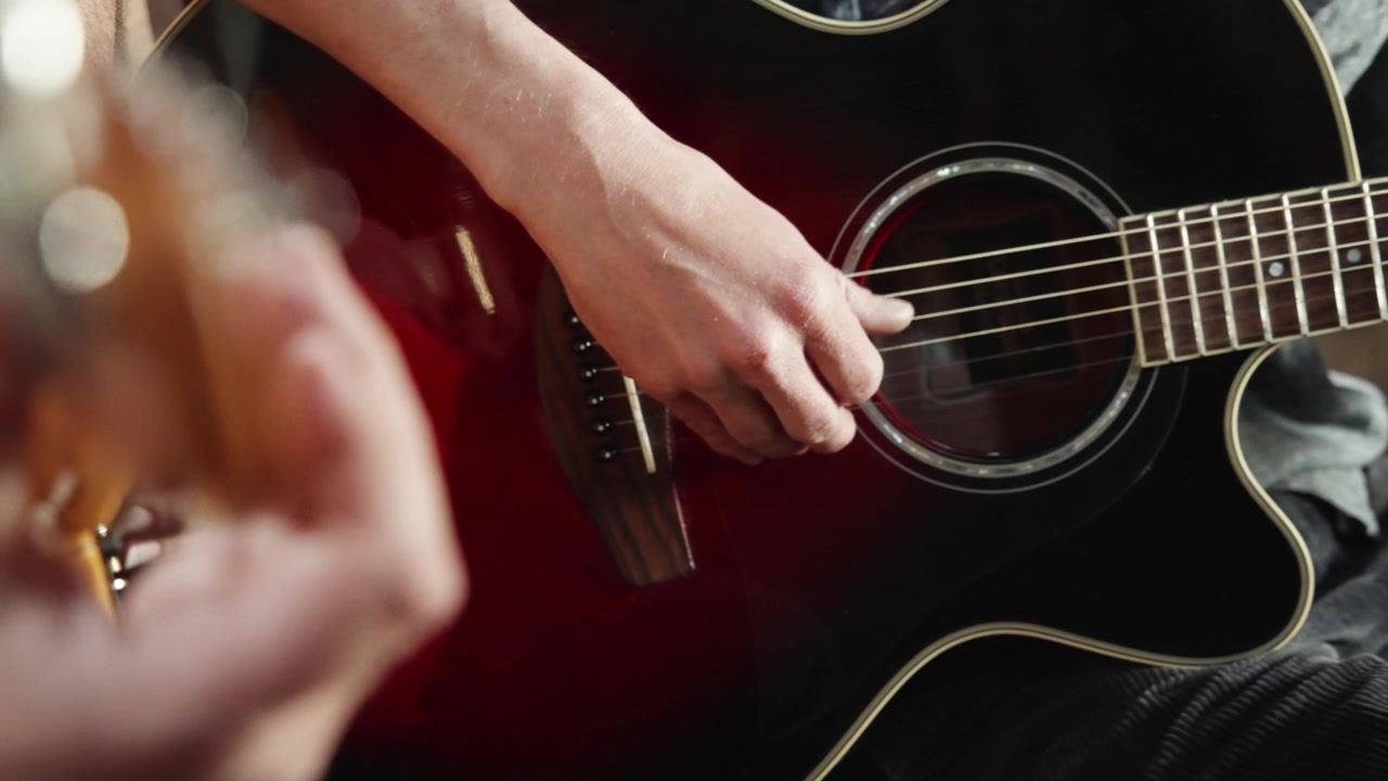 Zbliżenie na dłonie mężczyzny szarpiące struny gitary akustycznej w świetle reflektora, z dłonią drugiego gitarzysty na pierwszym planie
