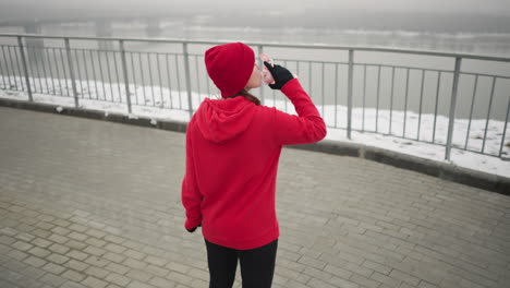 dama con capucha roja y gorra beba agua de botella rosa al aire libre durante un sereno día de invierno con suelo nevado, barandilla de hierro, río tranquilo y distante puente de niebla