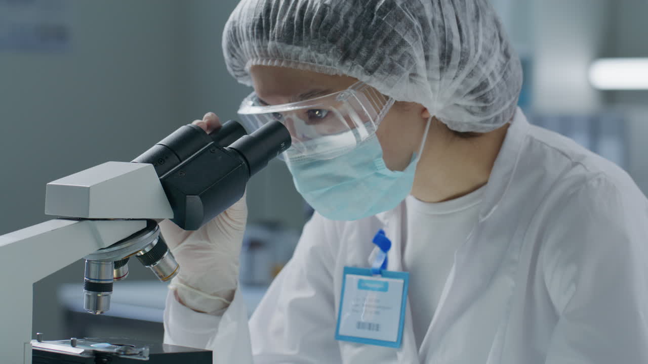 Masked Scientist Examining Liquid Sample under Microscope in Medical Laboratory