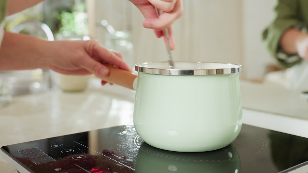 Close up of green pot on electric stove during cooking process with visible red control lights and reflective black glass surface while cook stirs food