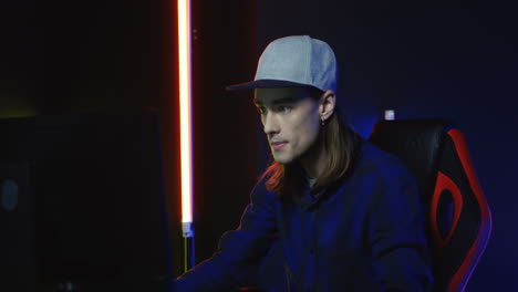 close up view of young serious man wearing cap and playing a game on the computer in a room with colorful neon lamps on the wall 3