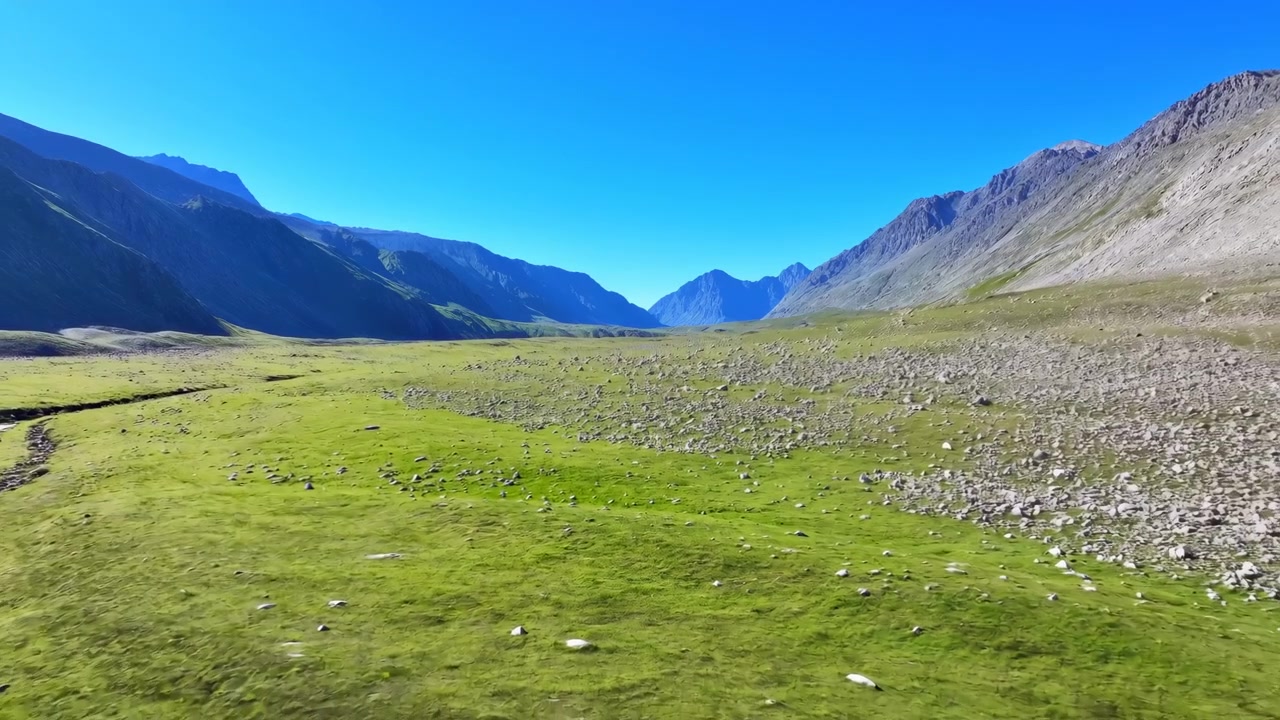 Rolig luftfoto over en vidstrakt høyfjellsdal som viser grønn eng, spredte steiner, en bred steinete slette, ulendte skråninger, fjerne topper, klar himmel og en rolig åpen villmark