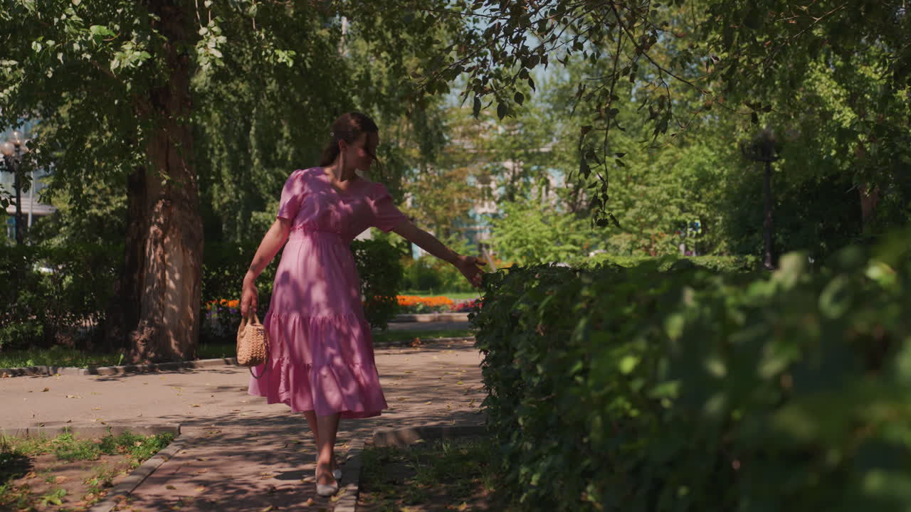 Mulher caucasiana passeando perto de uma cerca viva com um vestido rosa e bolsa, tocando as folhas e girando sob um dossel iluminado pelo sol, movimento rítmico lúdico pelo caminho do parque com canteiros de flores e árvores sombreadas