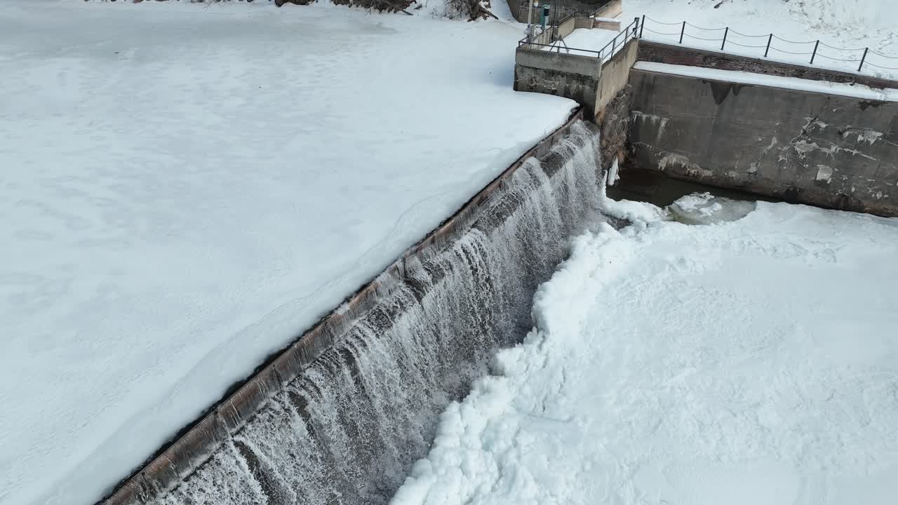 Aerial footage showing a drone moving backward to reveal a dam with water cascading over frozen structures, creating icy formations. Snow covers the landscape, enhancing the wintery view