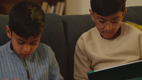 two young boys sitting on sofa at home playing games or streaming onto digital tablets 2