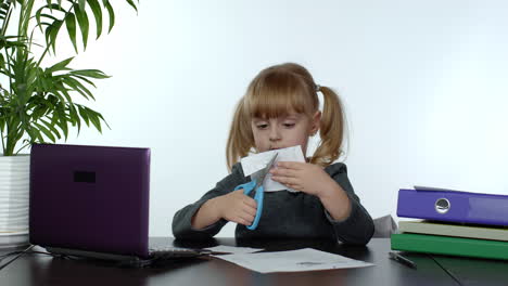 child schoolgirl learns lessons at home sitting at table cutting with scissors shapes out of paper