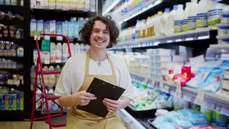 Portrait-of-a-happy-guy-with-curly-hair-in-a-yellow-apron-doing-inventory-and-holding-a-tablet-in-his-hands-in-the-dairy-section-of-a-supermarket