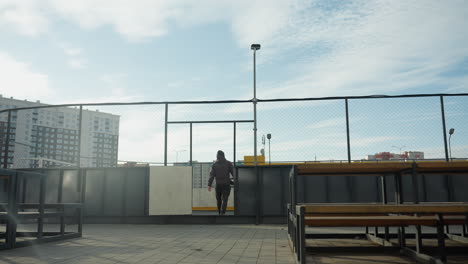 back view of person walking away from urban sports arena, carrying soccer ball, with vibrant yellow storage containers and city buildings in the background