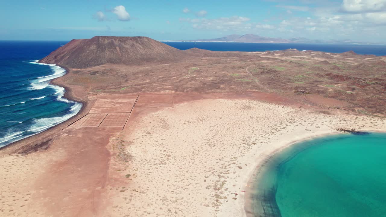 Drone view of Playa De La Concha De Lobos: vast sandy beach curving into turquoise waters, rugged terrain, volcanic mountain in the background, partly cloudy sky. Perfect for serene coastal footage.