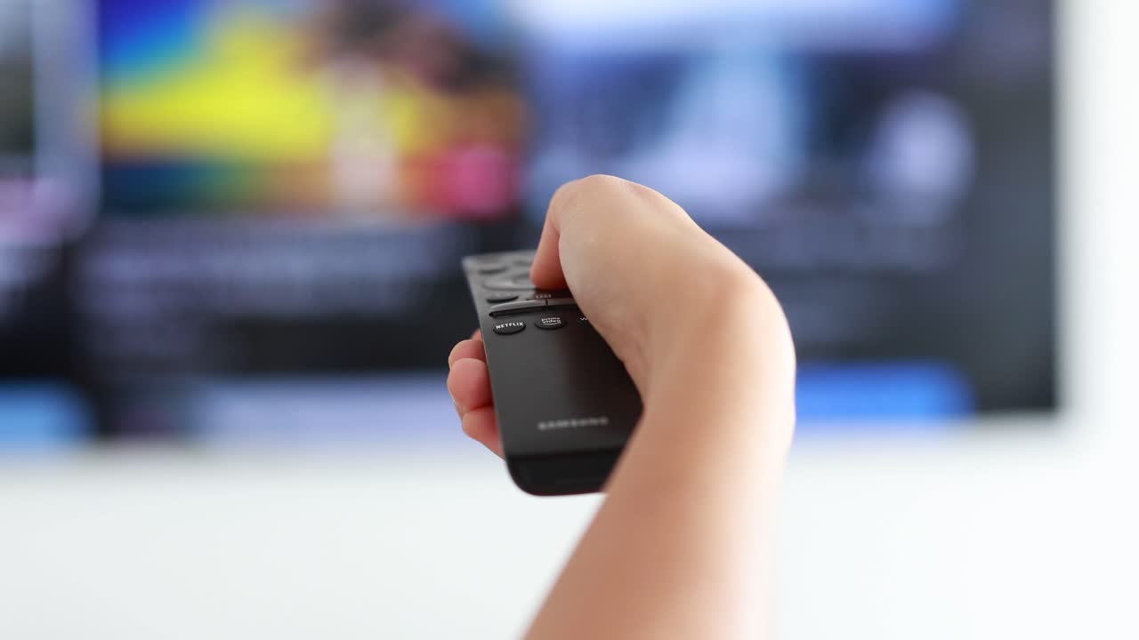 A person’s hand holds a remote control, pointing at a modern television while switching channels in a bright indoor living room with natural lighting