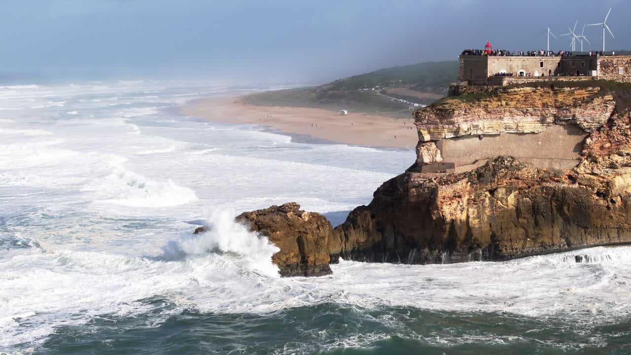 Drone aereo di onde enormi sulle scogliere di Nazaré, Portogallo. Faro di Nazaré e turbine eoliche. Tempesta atlantica