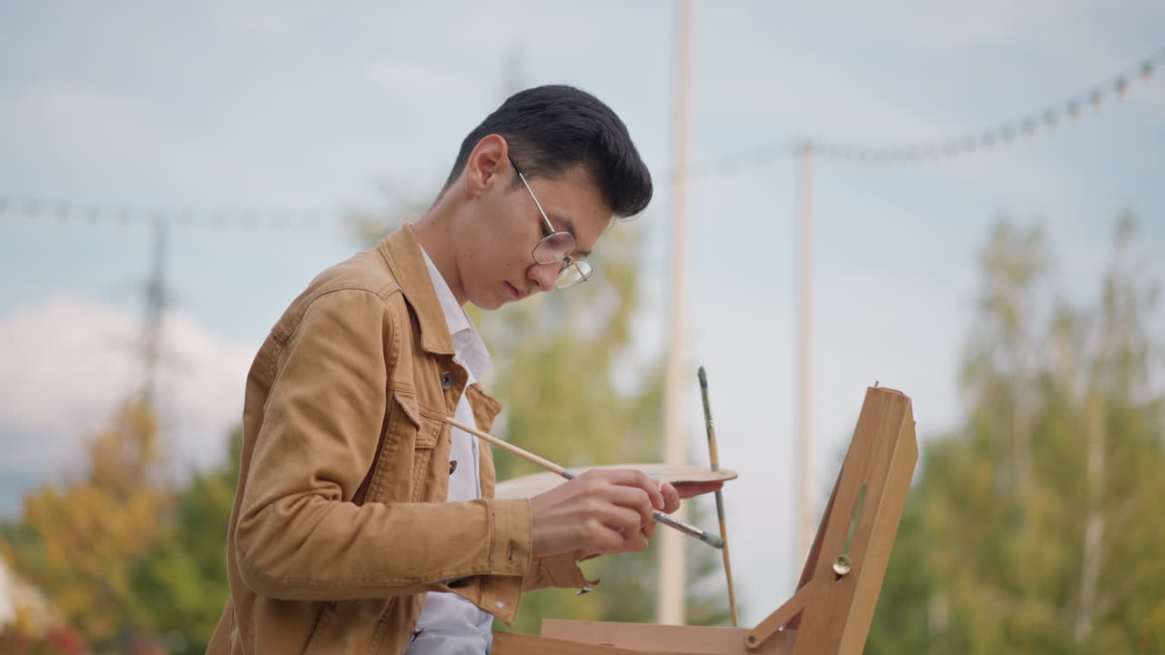 屋外のイーゼルで絵を描く若いアーティスト、木の箱のイーゼルとパレットを使った都会の公園でのスケッチセッション、集中した筆運びと色の混合、木々やスカイラインの観察、静かな創作活動と学習。